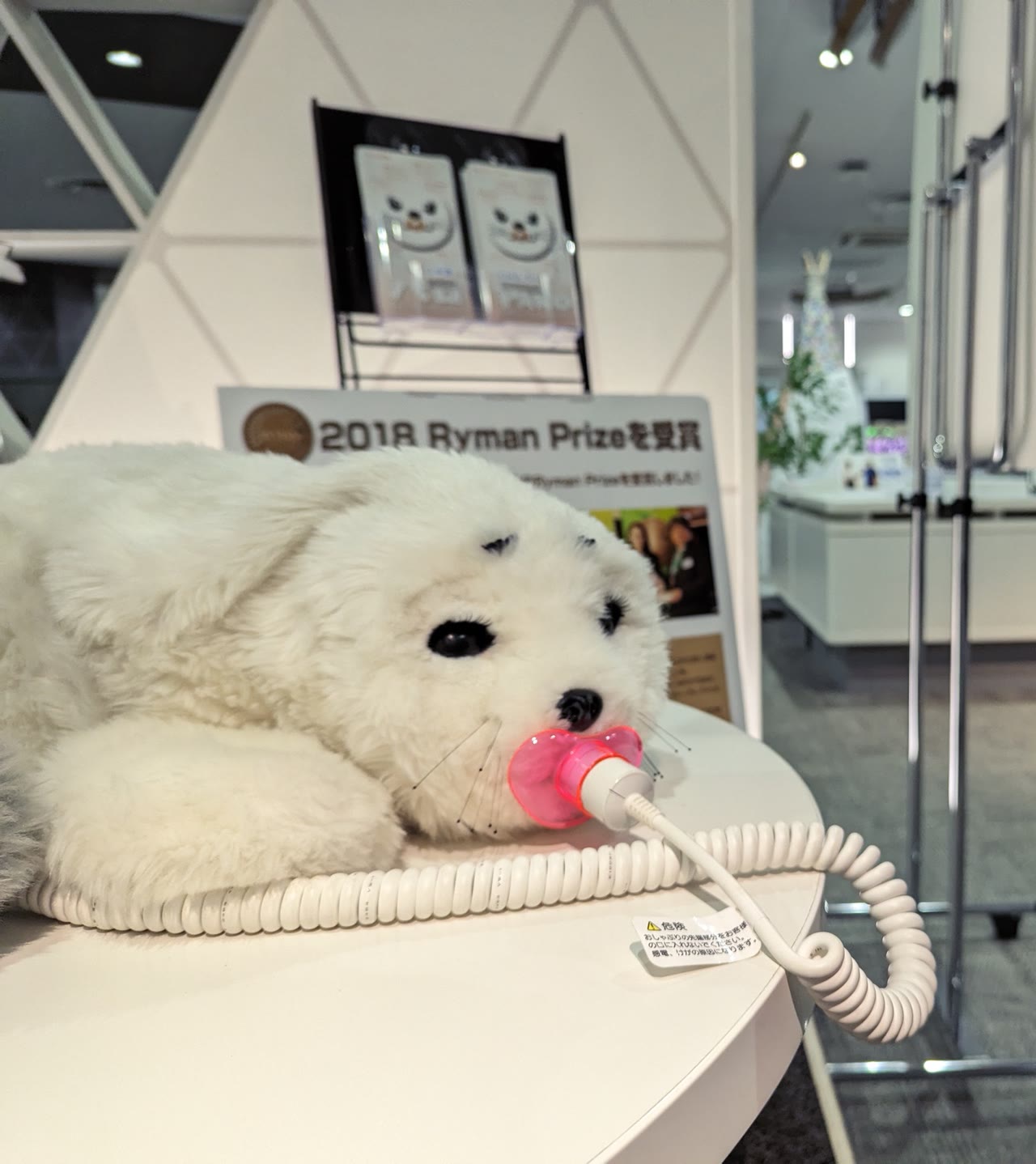 A white PARO therapeutic seal robot connected to a charging cable at an exhibition